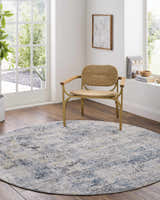 Bright room scene with a round Hyman Area Rug in gray, denim, and camel tones under a woven chair. Roomscene-Round