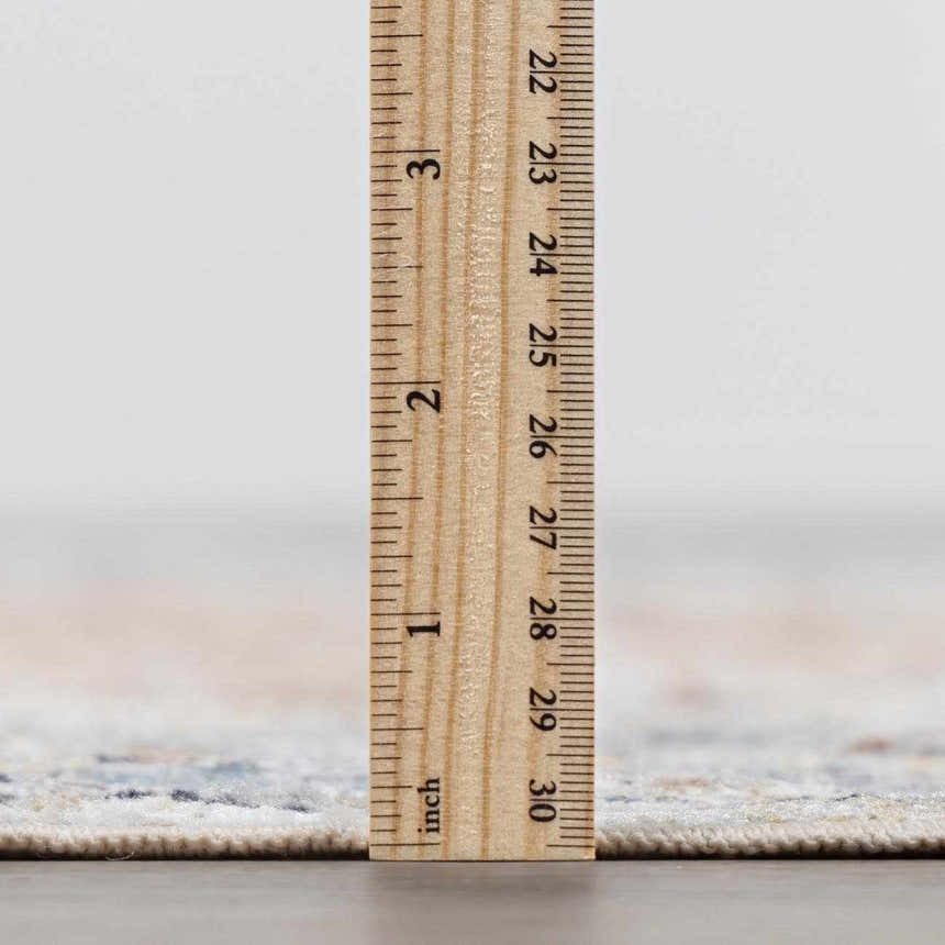 Close-up of the Emke Flat Pile Washable Rug showing the rug's pile height with a ruler. Bone, beige, and gray tones. Pile