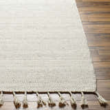 Close-up of the hand woven Abhay Beige Wool Area Rug with tassels on dark brown wood flooring. Front