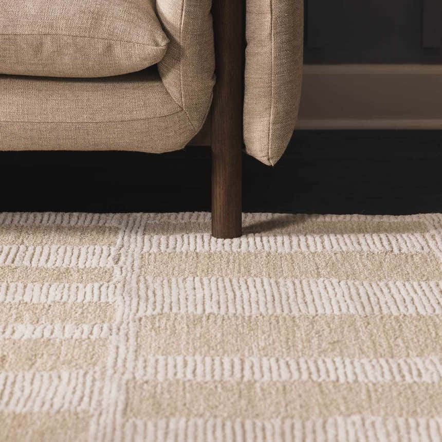 Close-up of the Teige Light Silver Area Rug featuring a textured, geometric pattern under a beige sofa. Styleshot