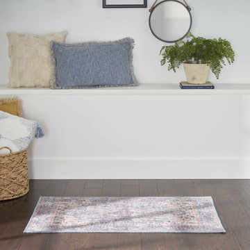 Entryway scene with blue pillows, plant, and Nourison New Concept Blue Multicolor Vintage Indoor Washable Area Rug on dark wood floor.