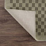Close-up of Gaiwan Olive Green Washable Area Rug showing checkerboard pattern, texture, and backing on wood floor. Fold