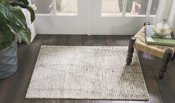 Entryway scene with a Nourison Ellora Stone rug, sandstone color, on dark wood floor near a woven stool and potted plant.