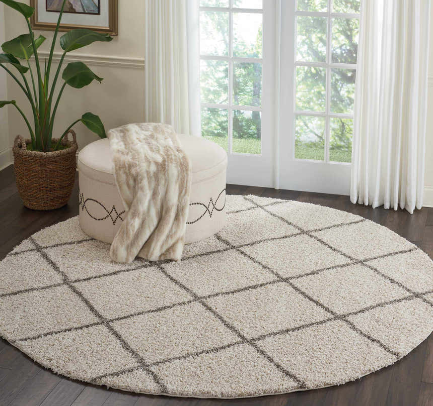 Living room scene with round cream shag Nourison Brisbane rug, ottoman, throw, plant, and natural light.