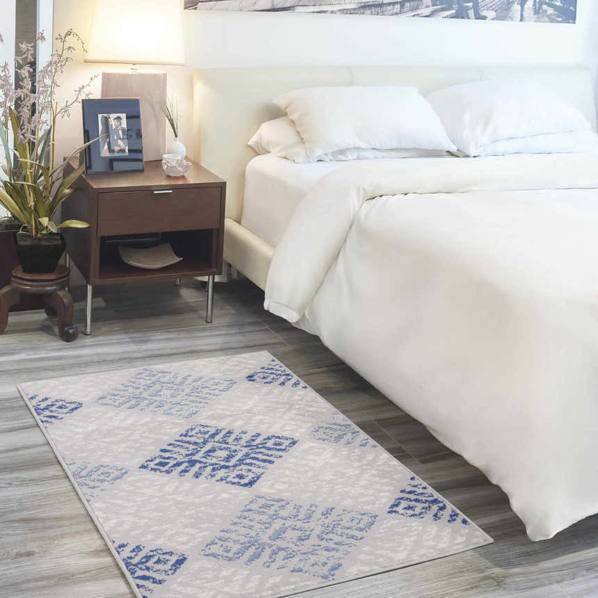 Bedroom scene with a Nourison Whimsicle Grey Blue Bohemian Indoor Area Rug, white bedding, and wood nightstand.