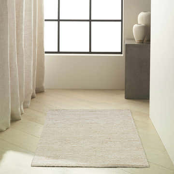 Minimalist room scene with a Calvin Klein Kathmandu Natural rug on light wood floors near curtains and a window.