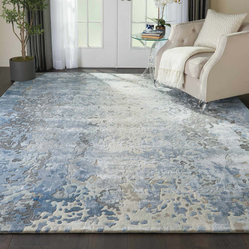 Living room scene with a Nourison Prismatic Denim rug, beige armchair, glass table, dark wood floors, and natural light.