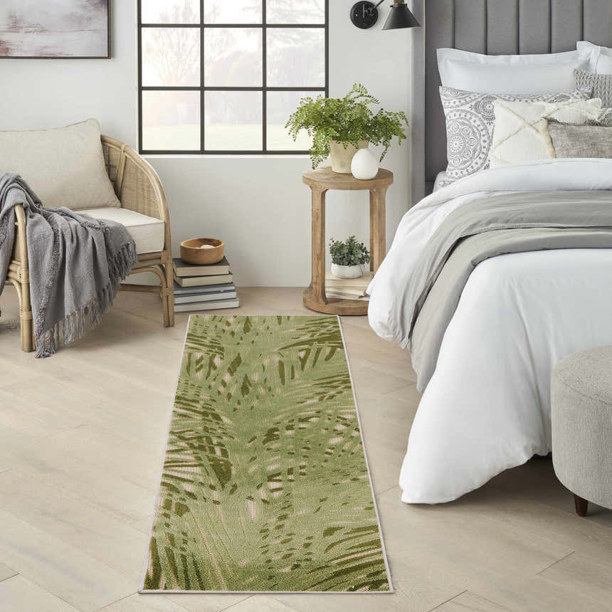 Bedroom scene with a Nourison Aloha Ivory Green Outdoor Area Rug, rattan chair, and bed with white linens.