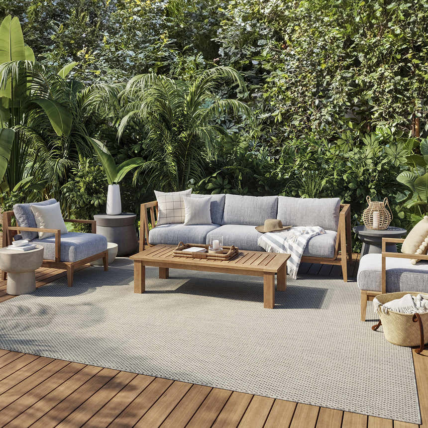 Outdoor patio scene with gray furniture and a Nourison Courtyard Ivory Silver rug on a wood deck surrounded by lush greenery.