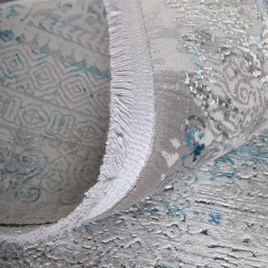 Close-up of the Feizy Cadiz rug showing its abstract blue, gray, and gold design and fringed edge. Fold