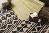 Styled Gratiot Area Rug scene: black and beige rug, rolled gray and yellow rug, trunk, yarn, and ottoman. Styleshot