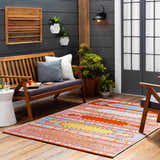Outdoor patio scene with a Gumberry Area Rug in brick red, bright red, and blue, styled with a wooden bench and plants. Roomscene-Rectangle