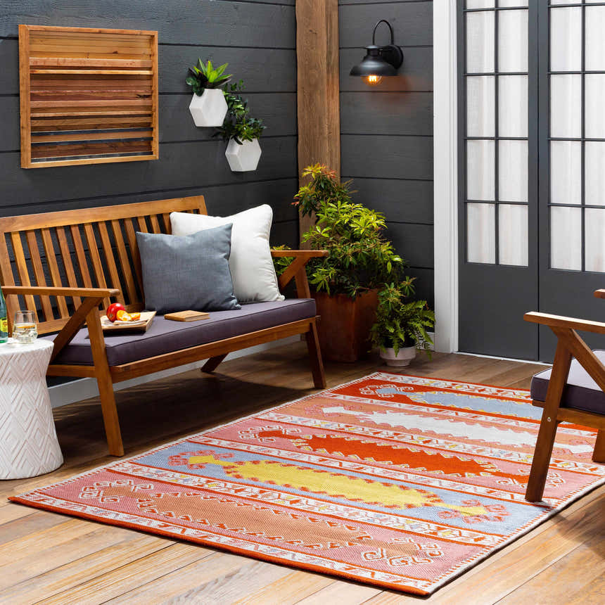 Outdoor patio scene with a Gumberry Area Rug in brick red, bright red, and blue, styled with a wooden bench and plants. Roomscene-Rectangle