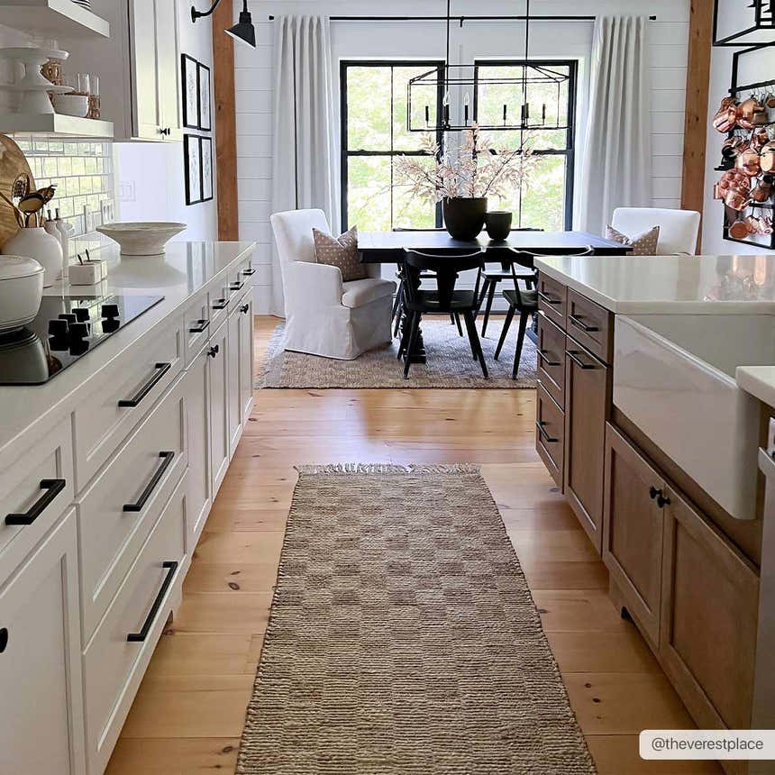 Farmhouse kitchen with a tan Ionut Checkered Jute Area Rug runner on wood floors, white cabinets, and dining area. Roomscene-Runner