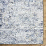 Close-up of the Hyman Area Rug. Features a contemporary design in camel, cloud gray, and denim hues with a distressed texture. Corner