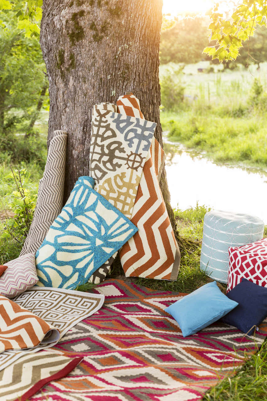 Outdoor scene with rolled Hollytree Area Rugs in cream, red, and blue, styled with pillows and poufs by a tree. Styleshot