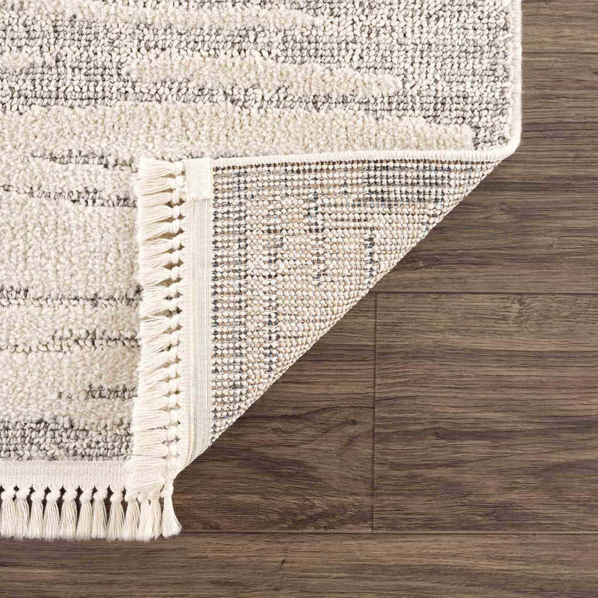 Close-up of the Lave Bone Area Rug showing its bone color, fringe, and woven construction on a dark wood floor. Fold