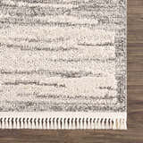 Close-up of the Lave Bone & Taupe Area Rug showing its textured, striped design and bone-colored fringe on a dark wood floor. Swatch