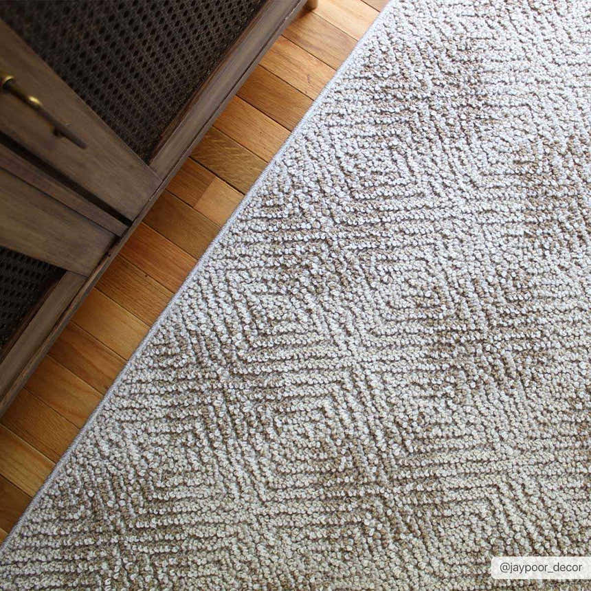 Close-up of the beige Broox Area Rug with a geometric diamond pattern on hardwood floors next to a wood cabinet. Roomscene-Rectangle