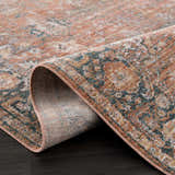 Close-up of the Mele Area Rug showing its traditional design, bone, burnt orange, and teal colors, and machine-woven texture. Fold