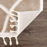 Close-up of the Azana Cream Area Rug showing geometric pattern, tassels, and the rug's backing on dark wood floor. Fold