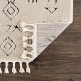 Close-up of the Thian Tribal Area Rug showing beige texture, black geometric pattern, white tassels, and rug backing. Fold