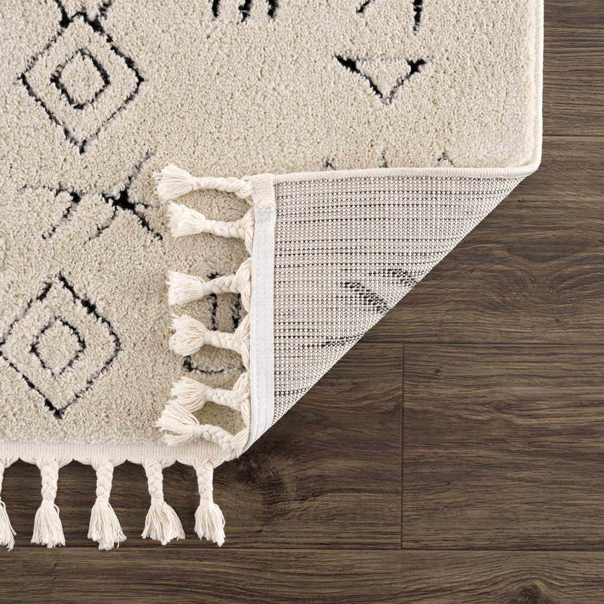 Close-up of the Thian Tribal Area Rug showing beige texture, black geometric pattern, white tassels, and rug backing. Fold