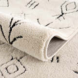 Close-up of the Thian Tribal Area Rug showing its beige color, plush texture, and black Moroccan-inspired geometric pattern. Fold