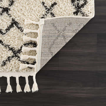 Close-up of the Godalming Plush Area Rug showing beige shag texture, charcoal pattern, and tassels on a dark wood floor. Fold