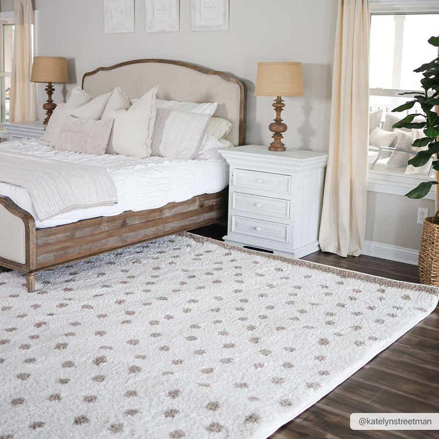 Bedroom scene with a Chaia Dotted Cream & Brown Plush Rug, wood bed frame, white nightstand, and neutral decor. Roomscene-Rectangle