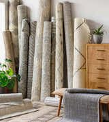 Rolled Beykoz Area Rugs in cream, beige, gray, and brown, displayed with a wood dresser, bench, and potted plant. Front
