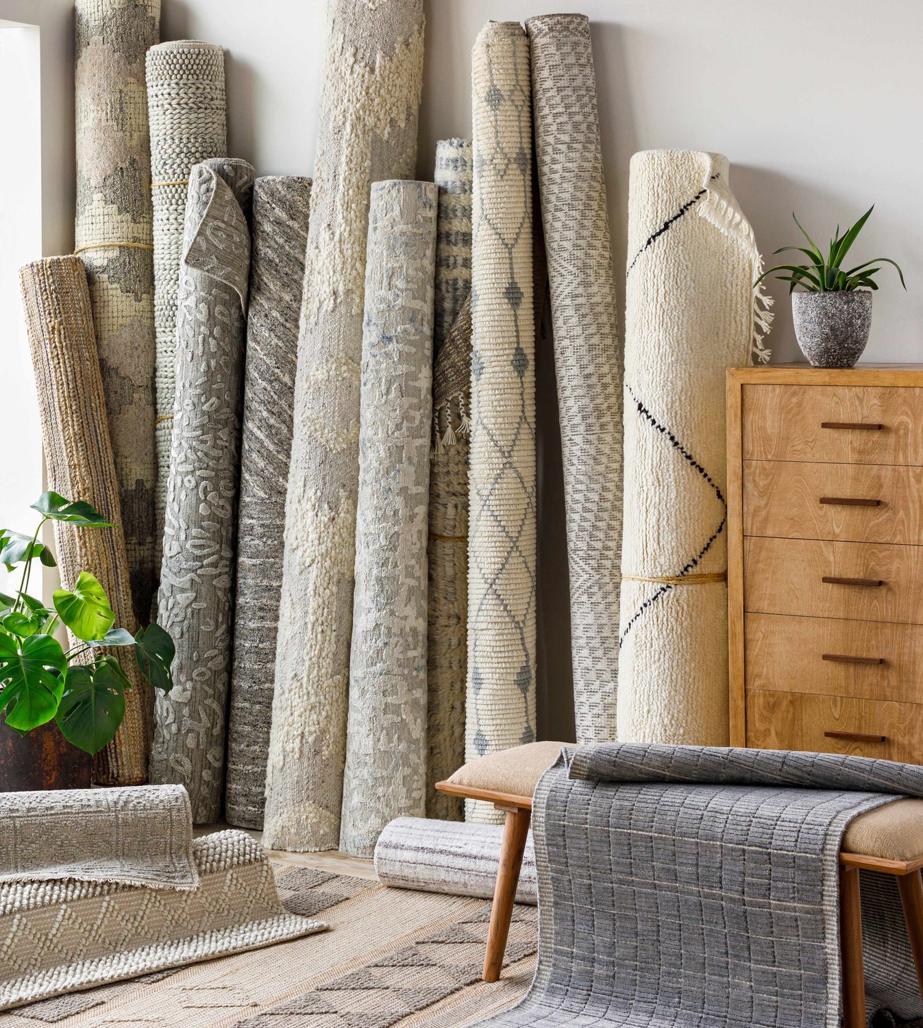 Rolled Beykoz Area Rugs in cream, beige, gray, and brown, displayed with a wood dresser, bench, and potted plant. Front