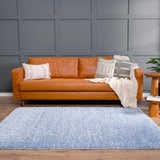 Living room scene with a Faina Solid Aqua Shag Rug, tan leather sofa, throw pillows, wood floors, and gray paneled wall. Roomscene-Rectangle