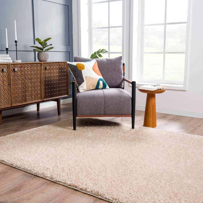 Living room scene with a Faina Solid Beige Shag rug, gray chair, wood cabinet, and ample natural light. Roomscene-Rectangle