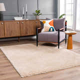 Living room scene with Faina Solid Beige Shag Washable Area Rug, gray walls, wood furniture, and a comfortable chair. Roomscene-Rectangle