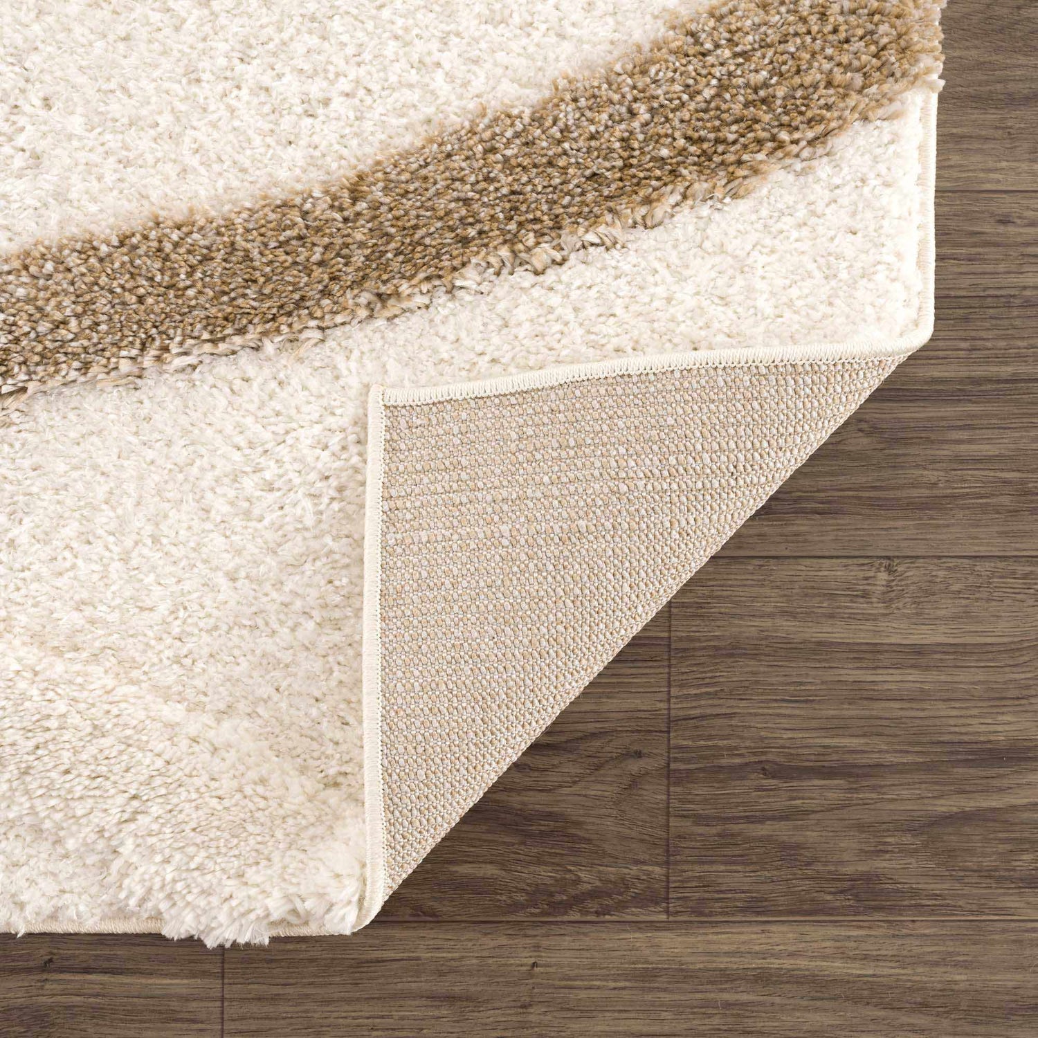 Close-up of the Espiye Area Rug showing the driftwood stripe, off-white shag texture, and backing on a dark wood floor. Fold