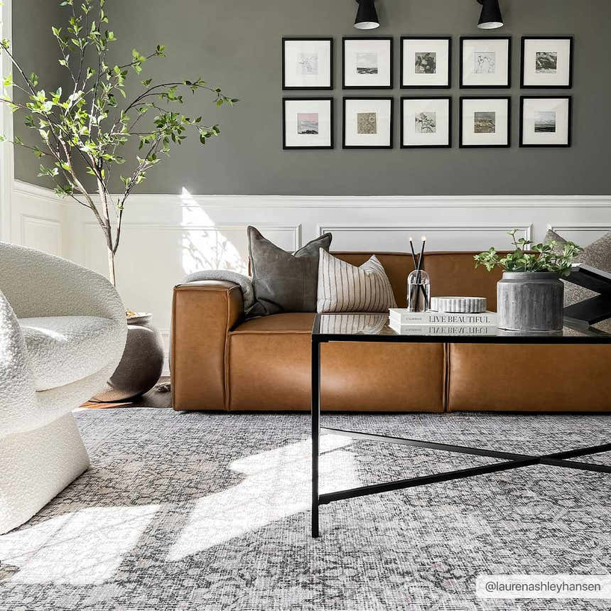 Living room scene with a gray Hedda Boutique Washable Rug, brown leather sofa, white chair, and black coffee table. Roomscene-Rectangle
