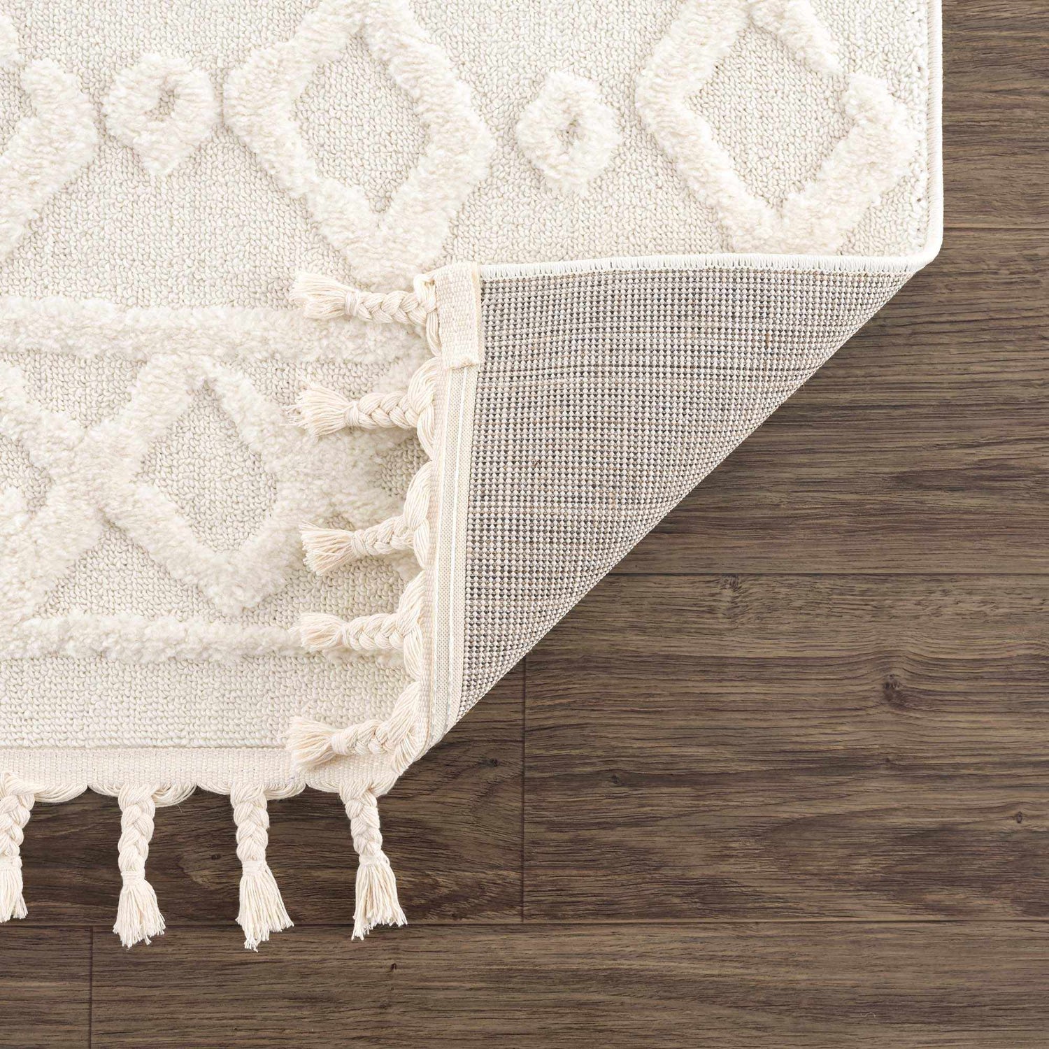 Close-up of the bone Gyundyukoru Area Rug showing geometric design, tassels, and machine woven construction on wood floor. Fold