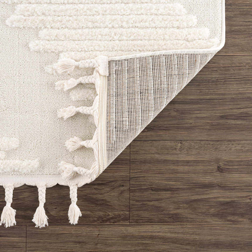 Close-up of the Palimbang Cream 3D Rug showing geometric textures, tassels, and the rug's backing on a wood floor. Fold