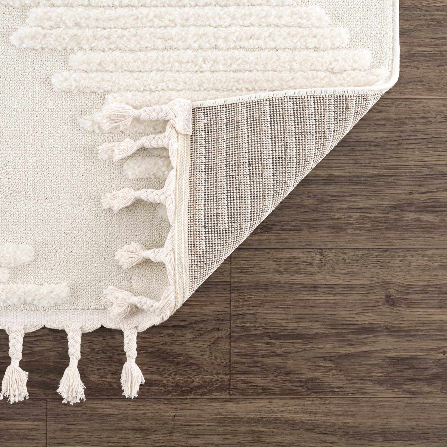 Close-up of the Palimbang Cream 3D Rug showing geometric textures, tassels, and the rug's backing on a wood floor. Fold