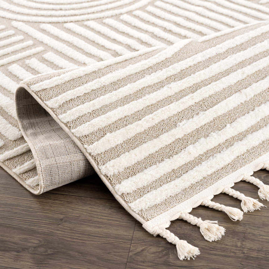 Close-up of the Moise Area Rug. Beige and cream rug with geometric and striped patterns, and decorative tassels. Fold