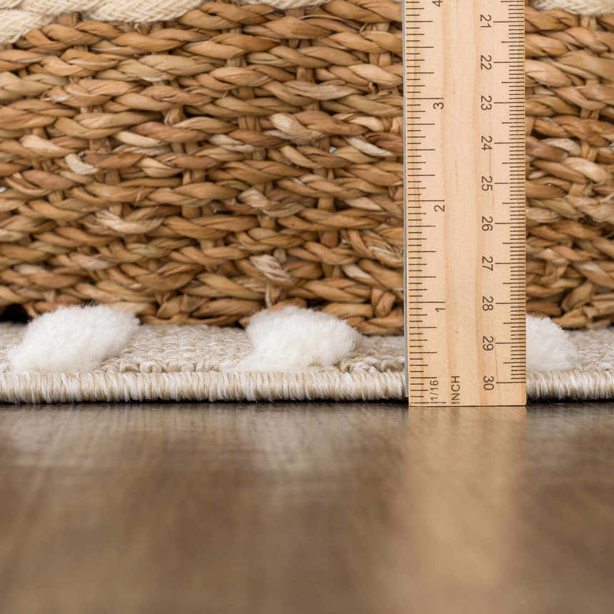 Close-up of the Moise Area Rug showing cream pom-pom details, beige woven texture, and ruler for scale. Pile
