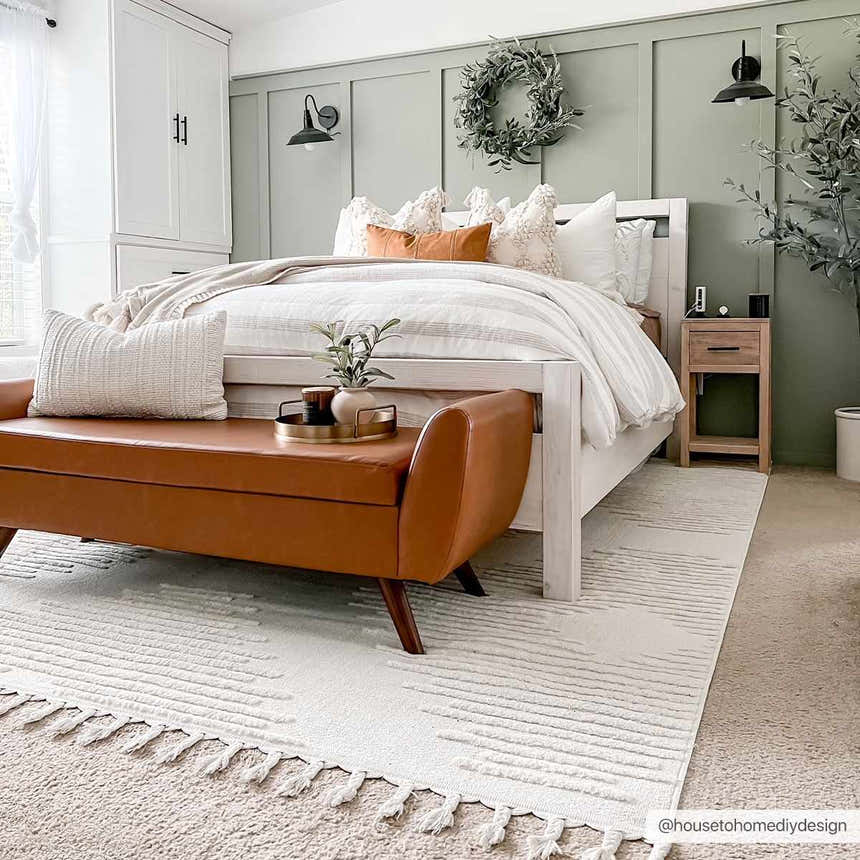 Bedroom scene with Palimbang Cream 3D Rug, white bedding, sage green walls, and a brown leather bench at the foot of the bed. Roomscene-Rectangle