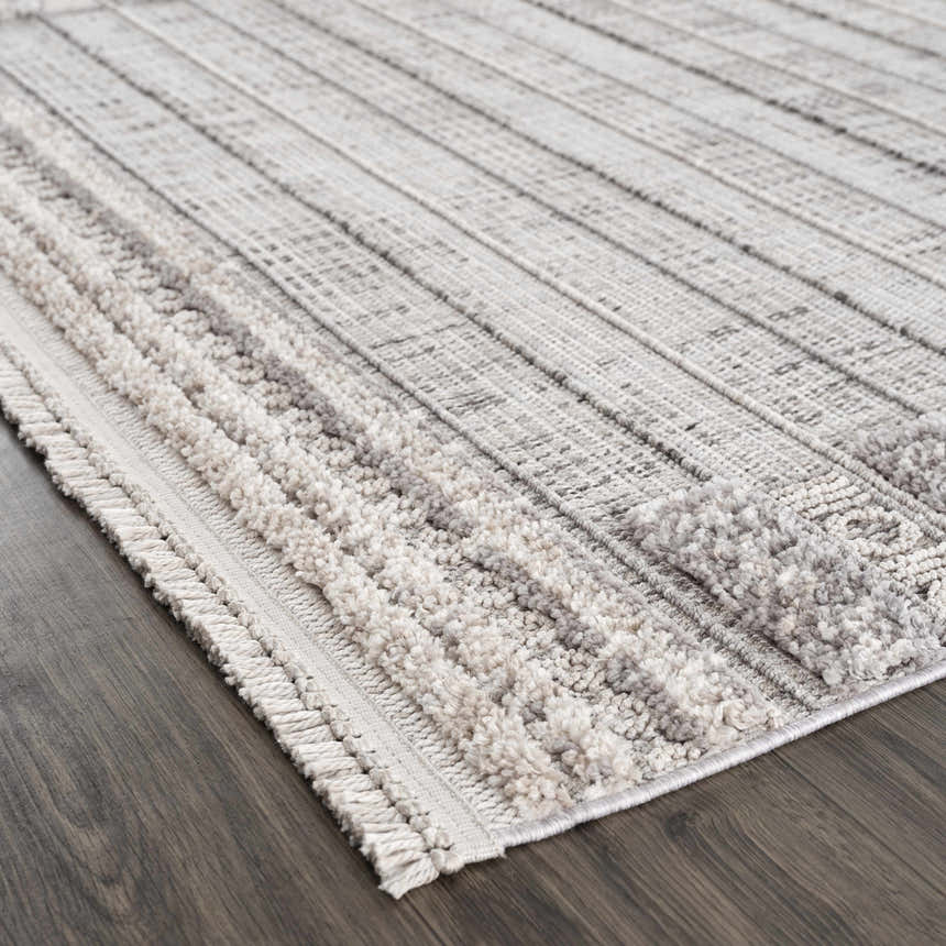 Close-up of the Naivasha Elegant High/Low Area Rug. Cream and charcoal rug with a textured, striped pattern and fringe detail. Corner