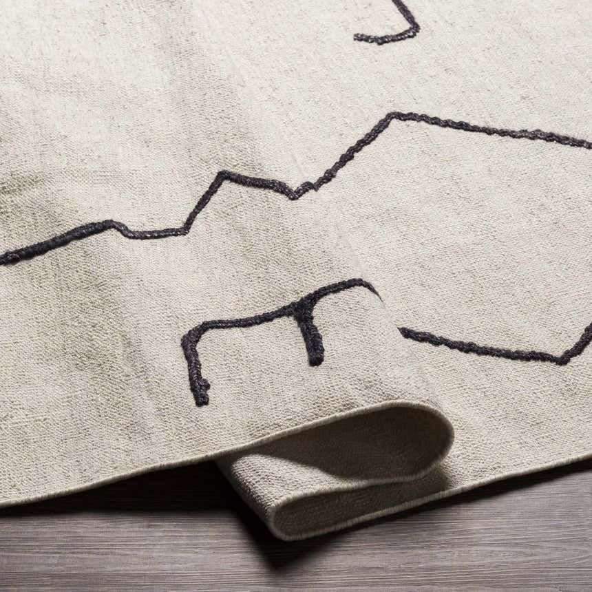 Close-up of the AISHA Area Rug. Hand-woven wool and viscose rug with a geometric black line pattern on a cream background. Fold