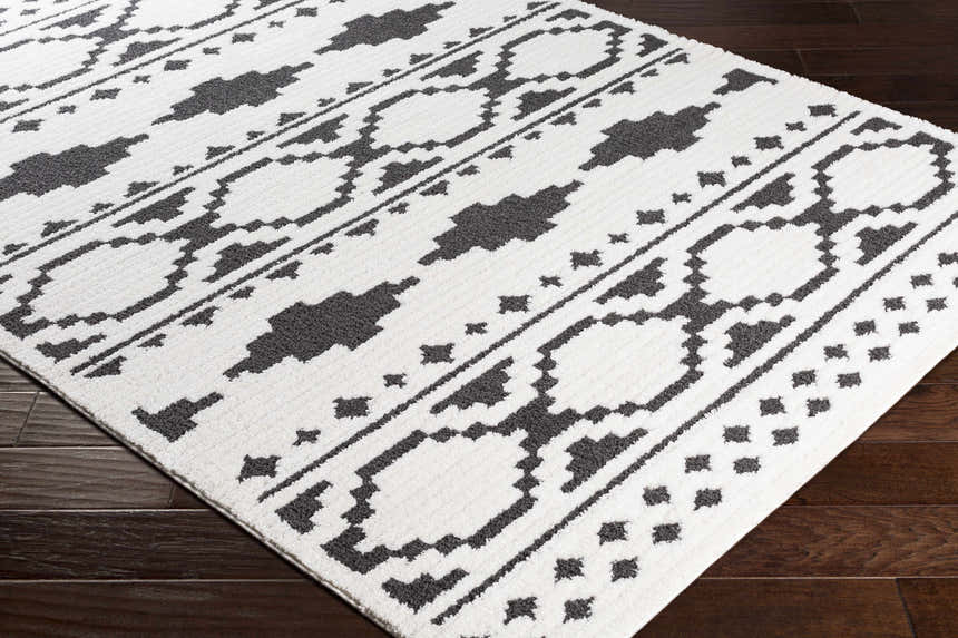 Close-up of the Amoret Area Rug, a machine-woven rug with a black and off-white geometric pattern on dark wood floors. Corner