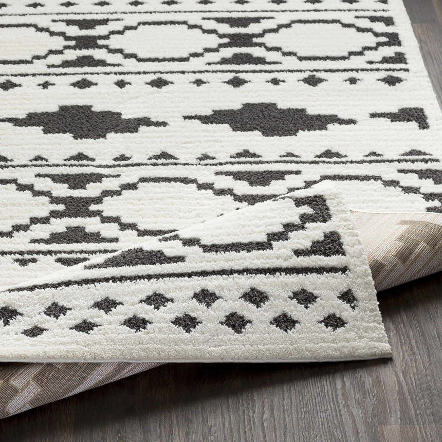 Close-up of the Amoret Area Rug showing its off-white base and charcoal geometric pattern. Machine woven texture visible. Fold