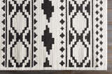 Close-up of the Amoret Area Rug. Black geometric pattern on off-white, machine woven texture on dark wood floor. Corner