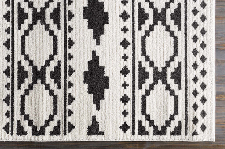 Close-up of the Amoret Area Rug. Black geometric pattern on off-white, machine woven texture on dark wood floor. Corner