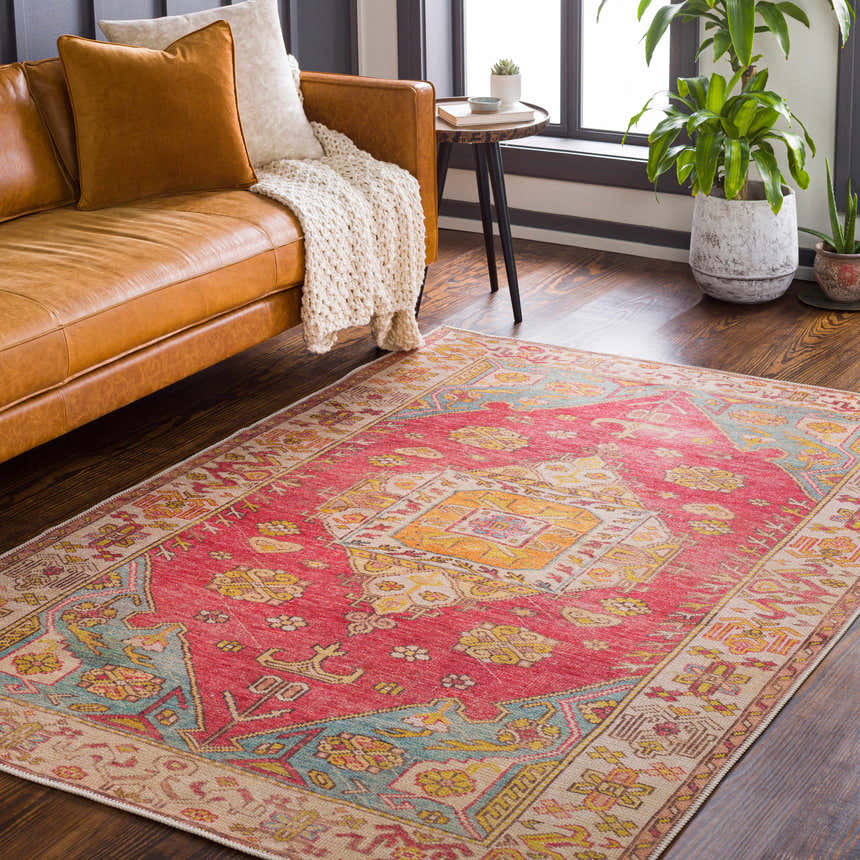 Living room scene with a red and gold Anopog Washable Area Rug, leather sofa, wood floors, and plants by the window. Roomscene-Rectangle
